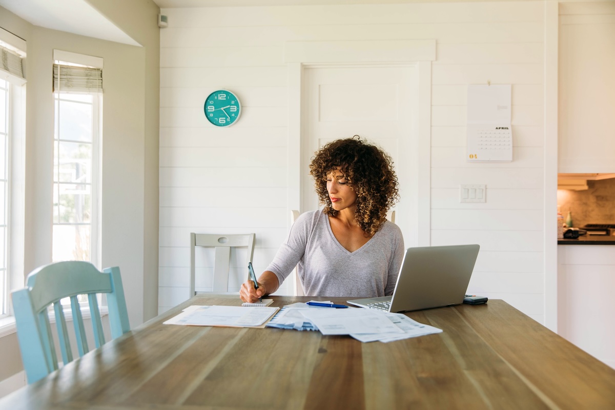 A woman sits at her kitchen table with her laptop surrounded by mail and paperwork