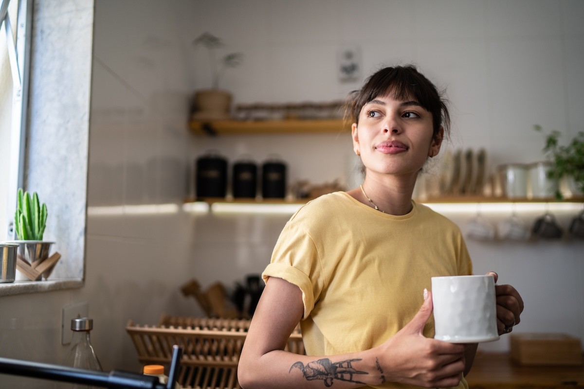 A woman in a yellow t-shirt stands in her kitchen drinking a cup of coffee.
