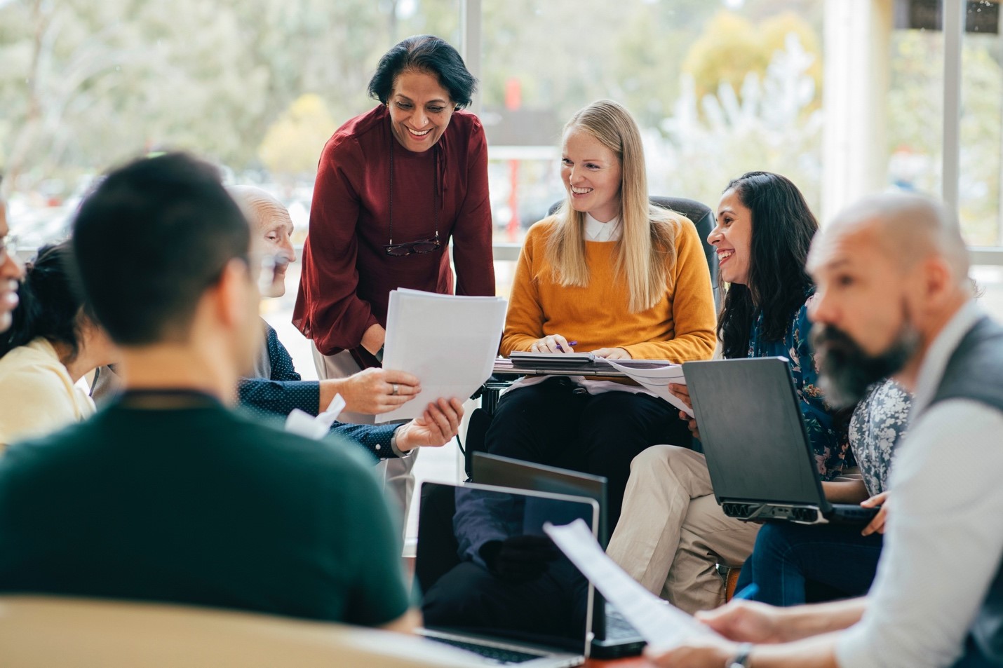Image of a group of 7 office workers talking and using laptops around a circle in an office 