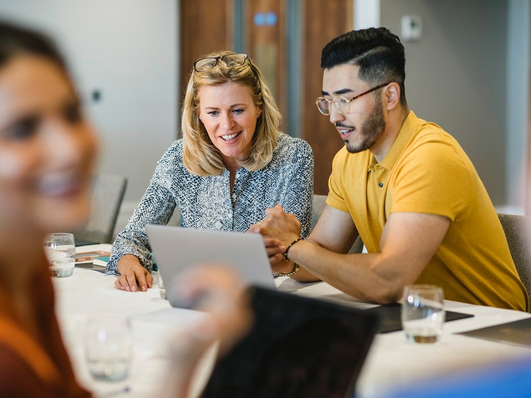 Picture of two coworkers in an office setting. A woman and a man sit side by side looking at benefits open enrollment information on a laptop. 