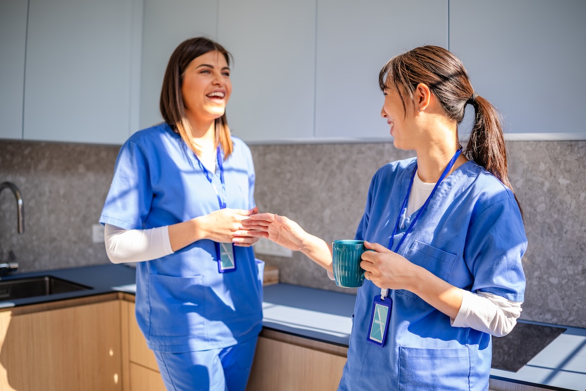 two nurses in blue scrubs standing in break room talking 