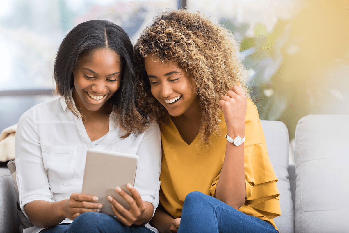 Two young women sit together on a sofa while looking at an iPad.