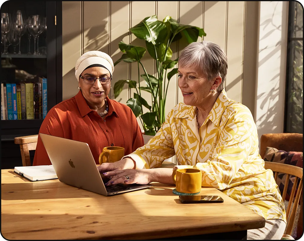 Two women sitting at a table using a laptop, each with a yellow mug, discussing something on the screen.