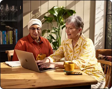 Two women sitting at a table using a laptop, each with a yellow mug, discussing something on the screen.