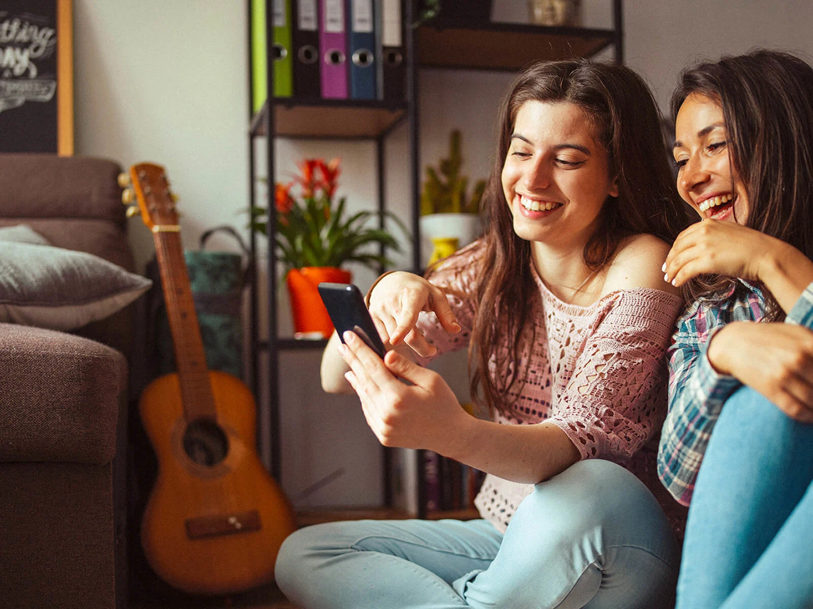 Two women enjoying time together while using a smartphone at home, representing staying connected with friends and family through digital communication.