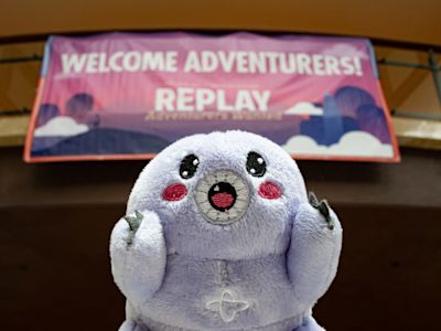 Light purple plush toy with embroidered face in front of pink Welcome Adventurers Replay sign