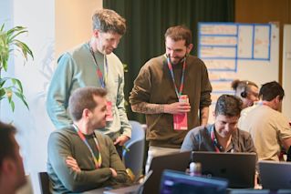 Conference attendees wearing lanyards collaborate around laptops in a room with a presentation board in the background.