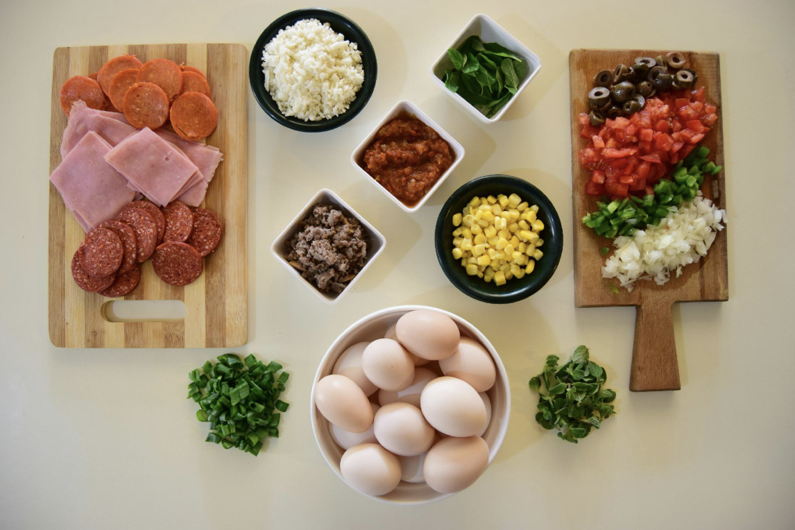 Prepped ingredients laid out in bowls and on cutting boards before any cooking starts: diced meat, cheese, herbs, tomatoes, olives, corn, and a bowl of eggs