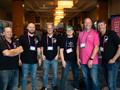 Six conference attendees wearing branded shirts and lanyards stand together in a hotel lobby with modern lighting.