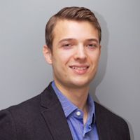 Young professional man smiling in black blazer and blue shirt against gray background, headshot portrait style.