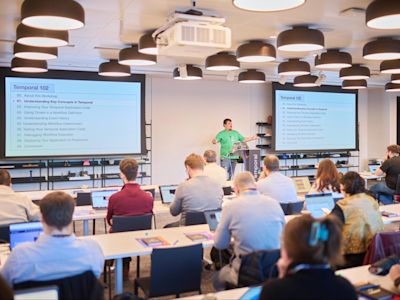 Instructor in green shirt presenting Temporal 102 workshop to seated audience in modern office with pendant lights and screens.