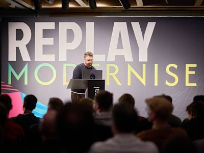 Speaker presenting at podium before audience with large REPLAY MODERNISE sign in white and gradient text on dark wall.