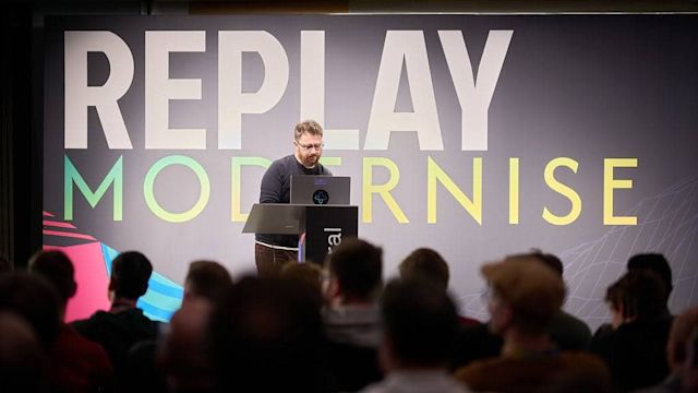 Speaker presenting at podium before audience with large REPLAY MODERNISE sign in white and gradient text on dark wall.