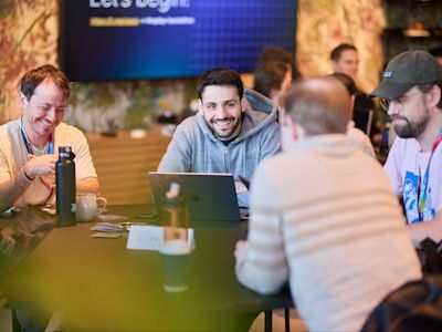 Group of smiling people collaborating at a table with laptops and water bottles at a casual tech event or workshop.