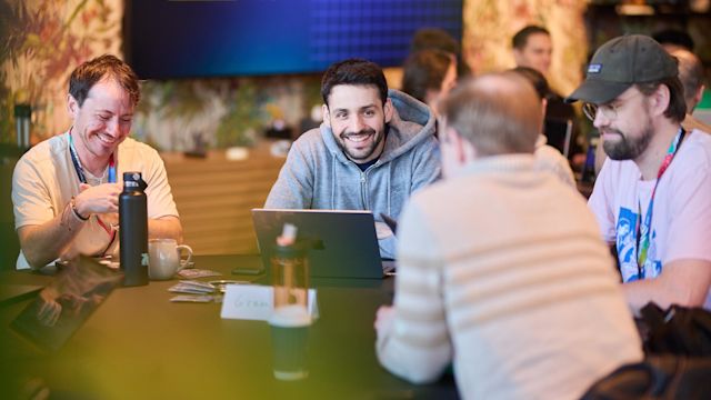 Group of smiling people collaborating at a table with laptops and water bottles at a casual tech event or workshop.
