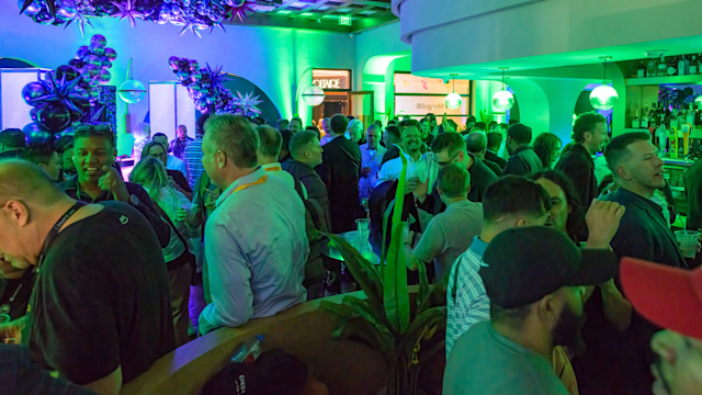 Crowded corporate event with attendees mingling under purple and green lighting with decorative balloons and displays.