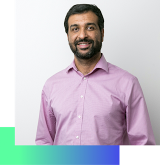 Professional headshot of a smiling man with dark hair and beard wearing a light purple button-down shirt on white background.