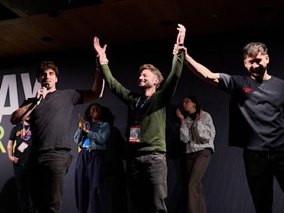 Three young men on stage celebrating with raised arms, center person in green sweater holds trophy aloft at awards ceremony.