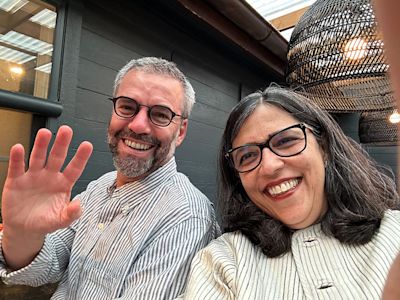 Two smiling people wearing glasses taking a selfie outdoors, man waving at camera, dark wood wall and wicker lamp behind them.