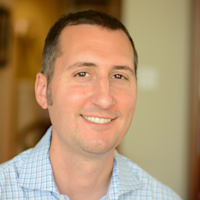 Professional headshot of a smiling man with short dark hair wearing a light blue checkered shirt against a blurred background.