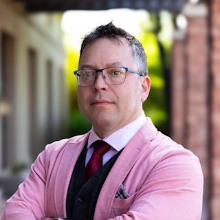 Professional man in pink suit jacket and black-framed glasses with arms crossed, standing outdoors with blurred background.