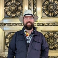 Bearded man in cap and navy jacket smiling in front of ornate yellow wall with decorative black medallions