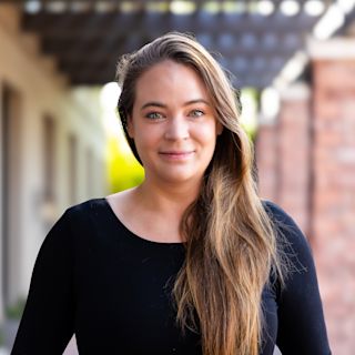 Woman with long brown hair wearing black top smiling at camera with blurred building background