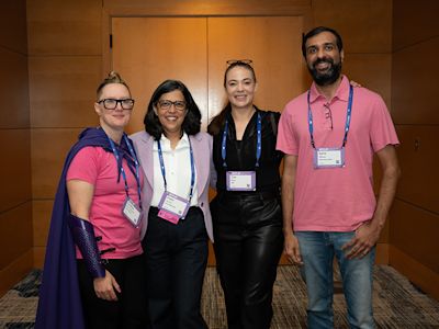 Four conference attendees wearing name badges smile together in front of wooden doors in a carpeted hallway.