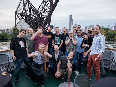 Group photo of diverse team on boat deck with Chicago skyline and industrial bridge in background under cloudy sky.