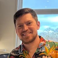 Man with short dark hair smiling at camera wearing colorful tropical floral shirt with window and trees in background