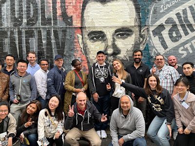 Diverse group of people posing together in front of a large painted mural featuring a portrait on a brick wall.