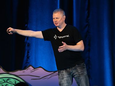 Man in black Temporal t-shirt gesturing while speaking on stage with blue curtain backdrop.