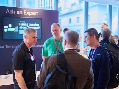 Attendees gather at an Ask an Expert booth with digital screen, conversing in a bright conference space with windows.
