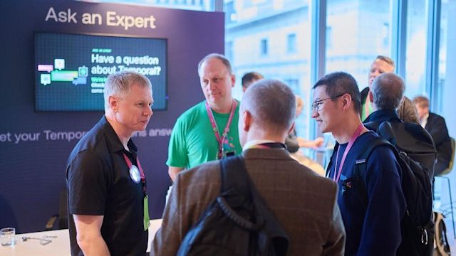 Attendees gather at an Ask an Expert booth with digital screen, conversing in a bright conference space with windows.
