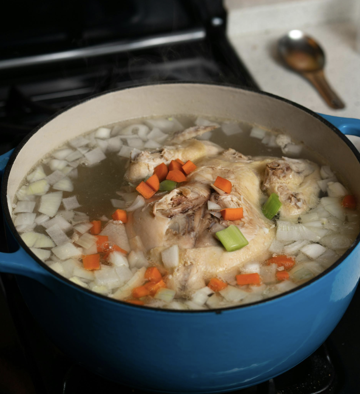 A blue enameled pot on a stove with chicken, carrots, celery and onion simmering down into stock