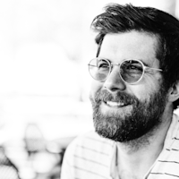Black and white portrait of a smiling bearded man wearing round glasses with styled hair against a light background.