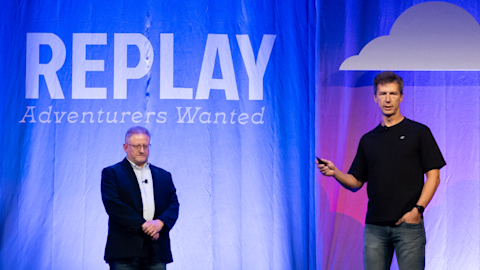 Two men on stage with blue backdrop displaying REPLAY Adventurers Wanted text and cloud graphic with purple lighting.