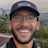 Smiling man wearing Patagonia cap and glasses taking selfie with lush green mountains and cloudy sky in background.