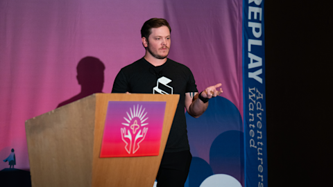 Man in black shirt presenting at wooden podium with colorful logo against purple backdrop and blue banner.