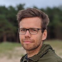 Man with brown hair and black-framed glasses wearing an olive green jacket outdoors with trees in the background.