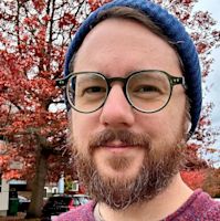 Man with beard and round glasses wearing blue beanie outdoors with red autumn foliage in background.