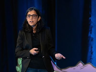 Woman with glasses and black blazer speaking at event, gesturing with hands against blue backdrop.
