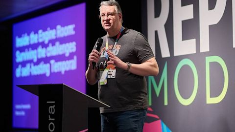 Speaker with microphone presenting at conference with purple slide screen and large text backdrop on stage.