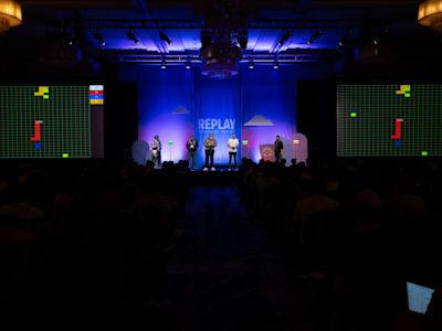 Stage presentation with five people under blue lighting, flanked by large screens displaying Tetris-style game graphics.