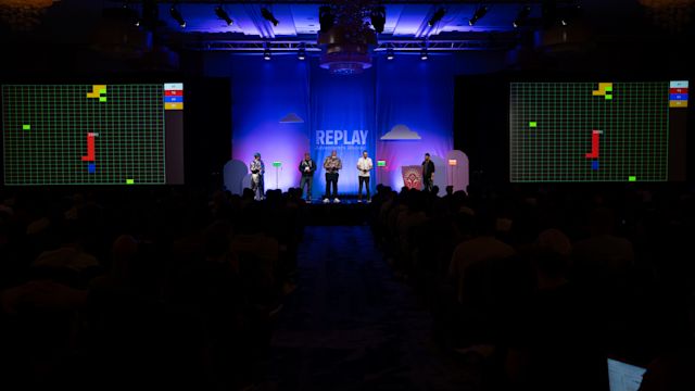Stage presentation with five people under blue lighting, flanked by large screens displaying Tetris-style game graphics.