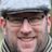 Smiling man with beard wearing black-framed glasses and gray herringbone flat cap with striped scarf