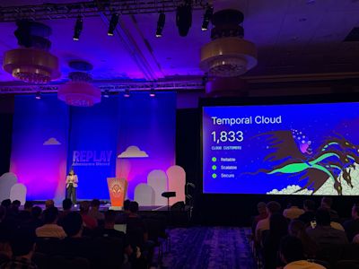 Conference presentation showing Temporal Cloud slide with 1,833 customers, speaker on stage, purple lighting, audience seated.