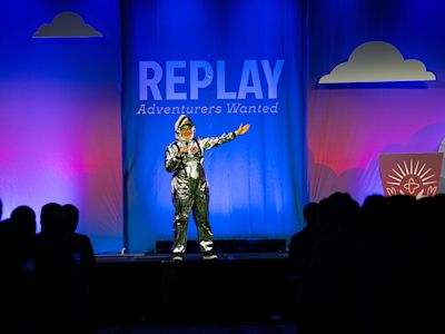 Person in silver astronaut costume on stage with blue backdrop reading REPLAY Adventurers Wanted, audience silhouettes visible.