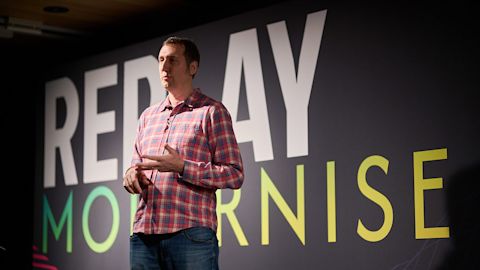Man in red plaid shirt presenting on stage with large text reading REPLAY MODERNISE projected behind him.