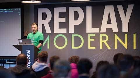 Speaker in green shirt presenting at podium with laptop before audience, large Replay Moderni sign on wall behind.